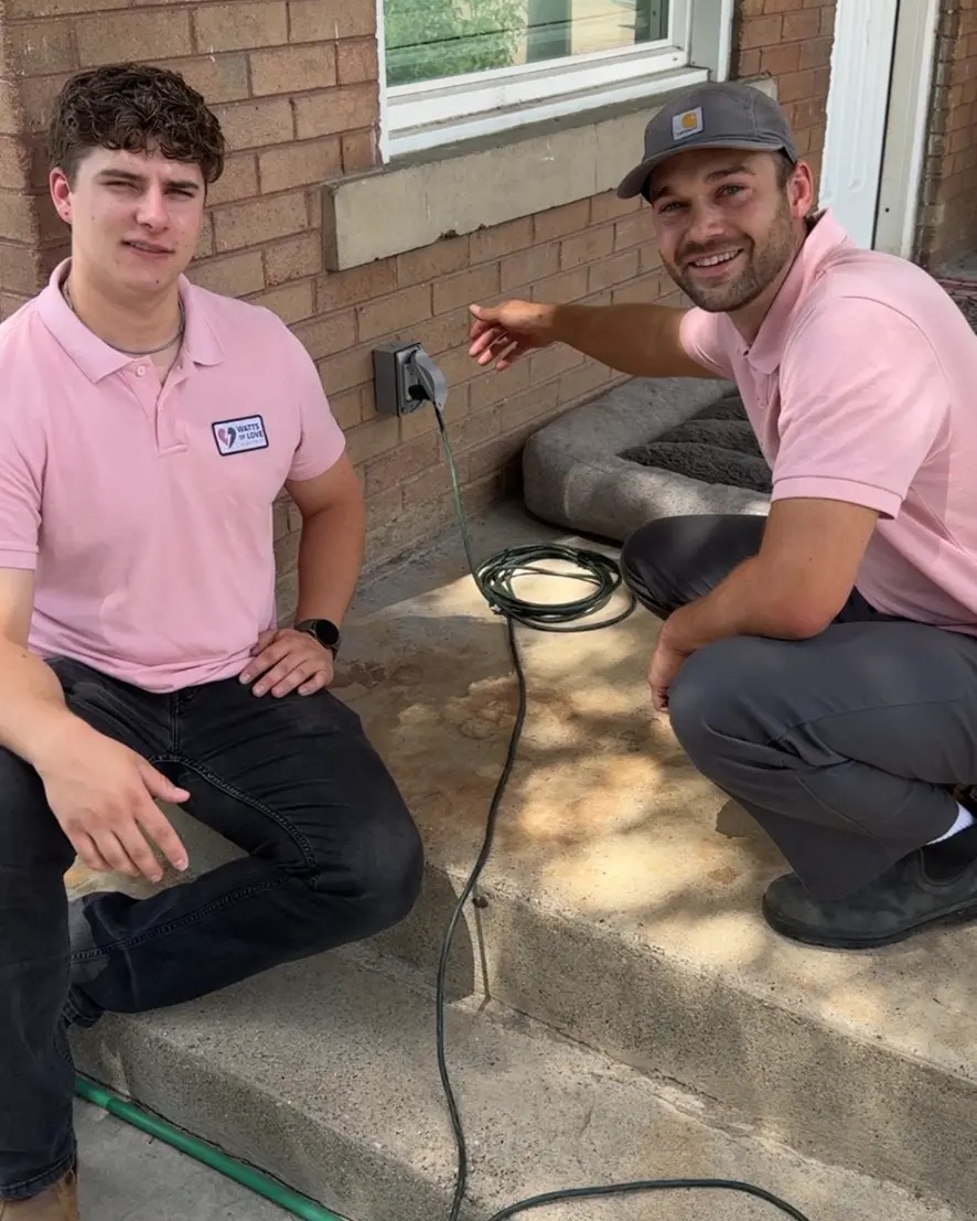 Two Watts of Love Electric technicians on the steps of a brick home, one pointing to an outdoor electrical outlet.